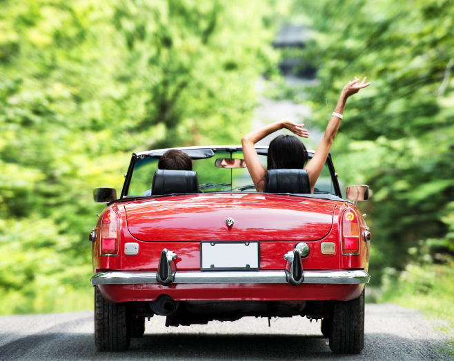 Two people driving/riding in a red convertible down a street lined with trees.  Passenger has their arms in the air.